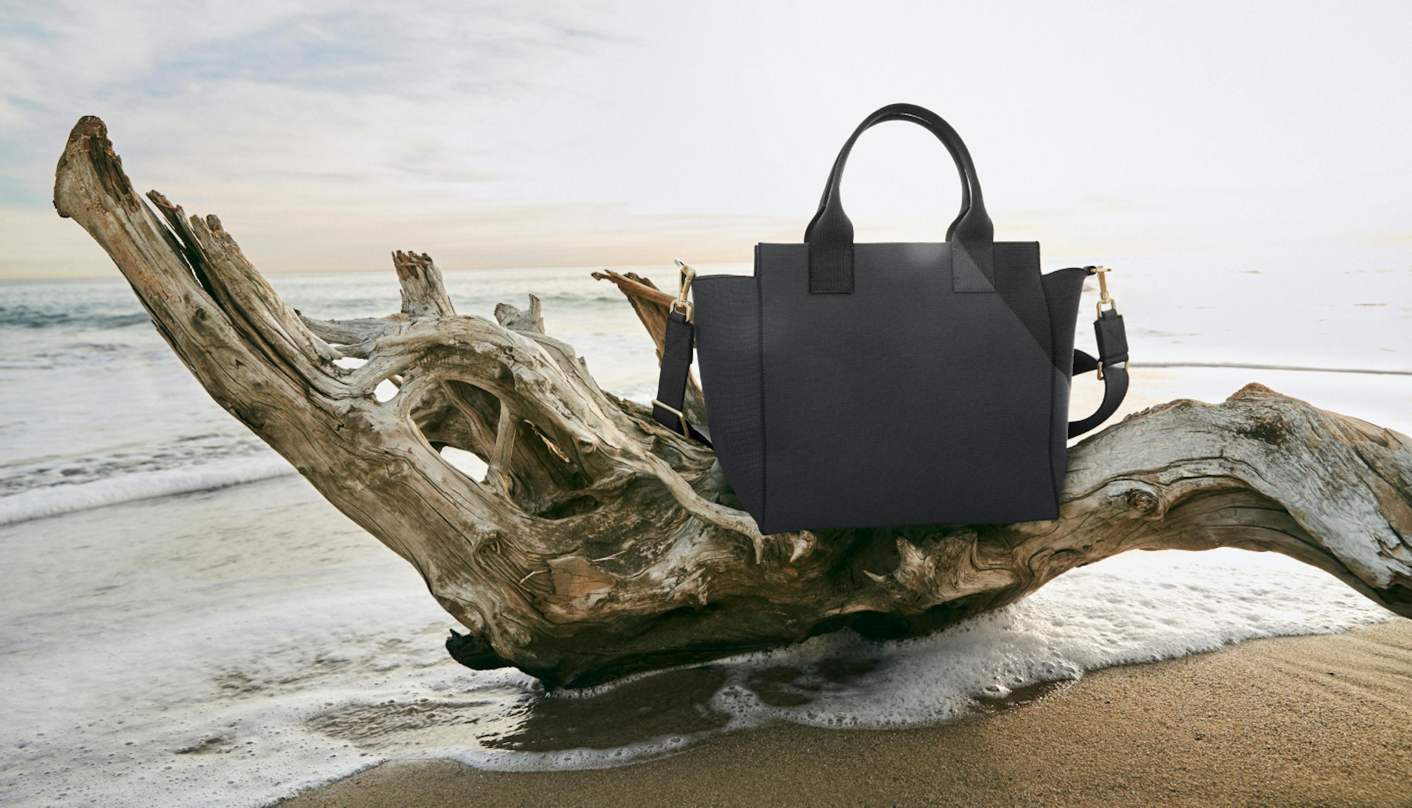 The Handbag in Slate Black sitting on a log by the ocean.