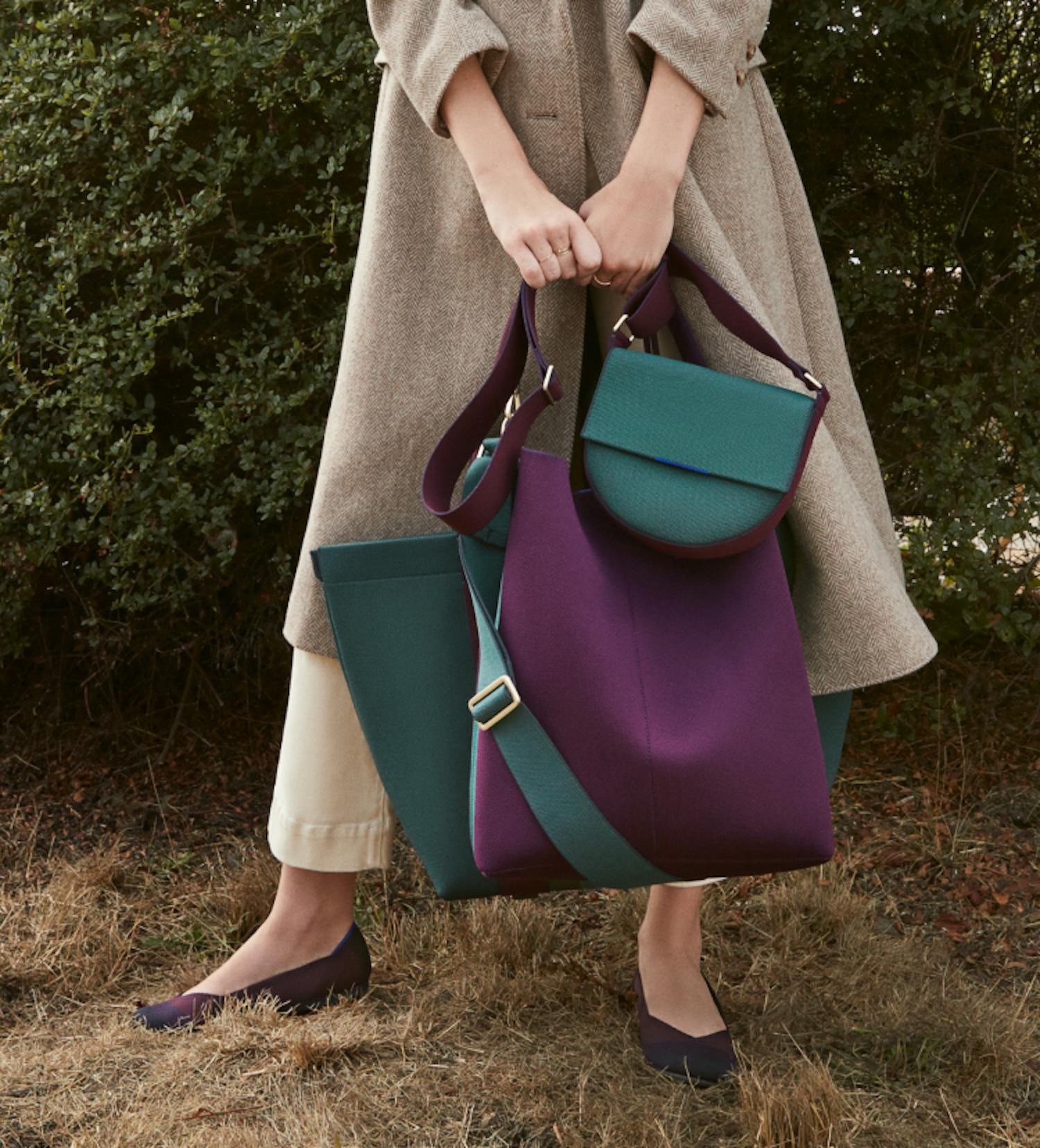 A model holding The Saddle Bag in Deep Spruce, The Bucket Bag in Dark Plum and The Essential Tote in Deep Spruce while wearing The Flat in Blackberry Captoe.