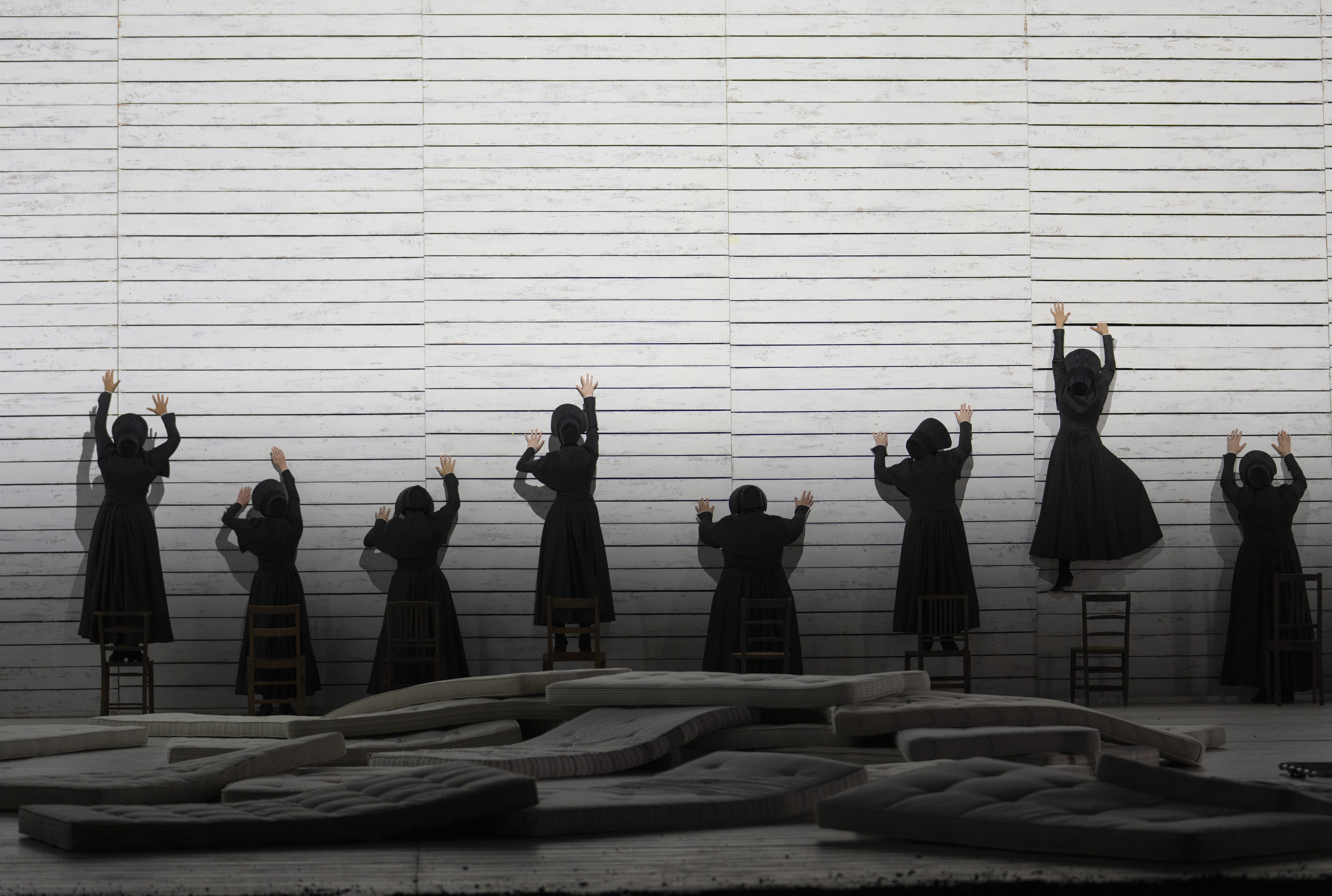 A group of women dressed in long black dresses attempt to climb up a sheer white wall. They have their backs to the camera.
