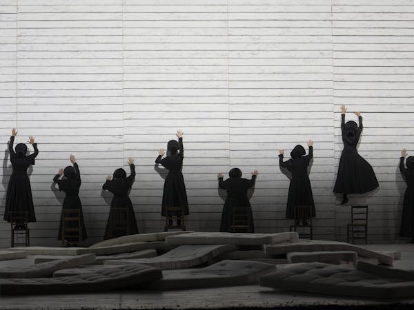 A group of women dressed in long black dresses attempt to climb up a sheer white wall. They have their backs to the camera.