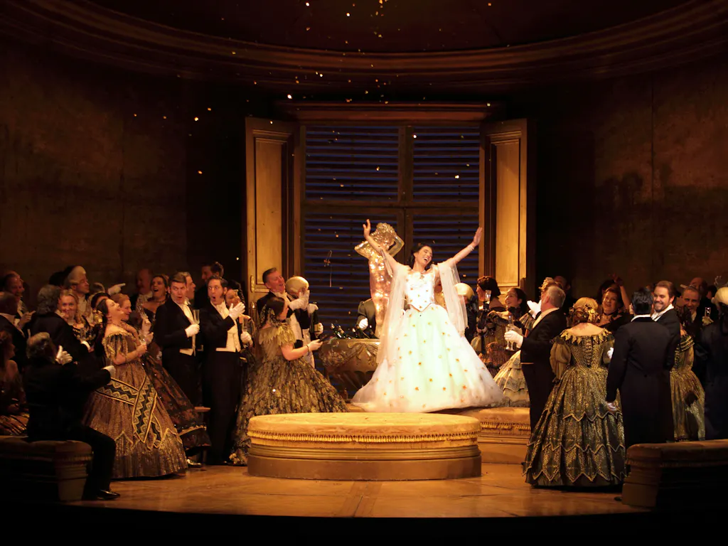 Violetta in a sparkling white dress stands in the centre of a golden room, bathed in warm light. Men and woman in formal suits and ballgowns look towards her as she stands with her arms stretched upwards.
