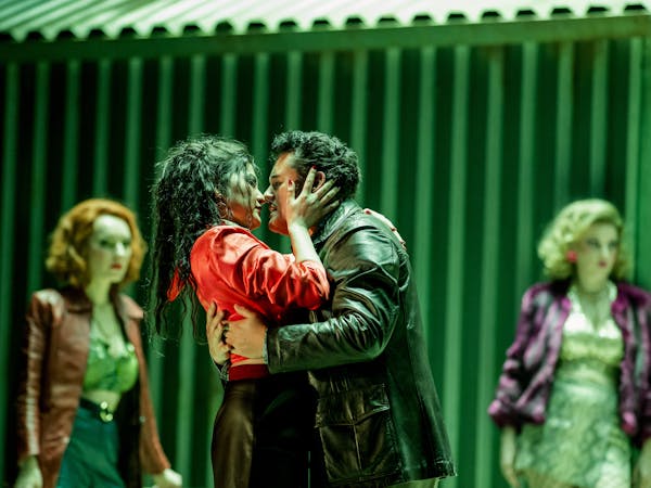 An opera singer with long black hair wearing red holds the head of another opera singer wearing a leather jacket in a passionate embrace. Two performers stand behind them looking on.