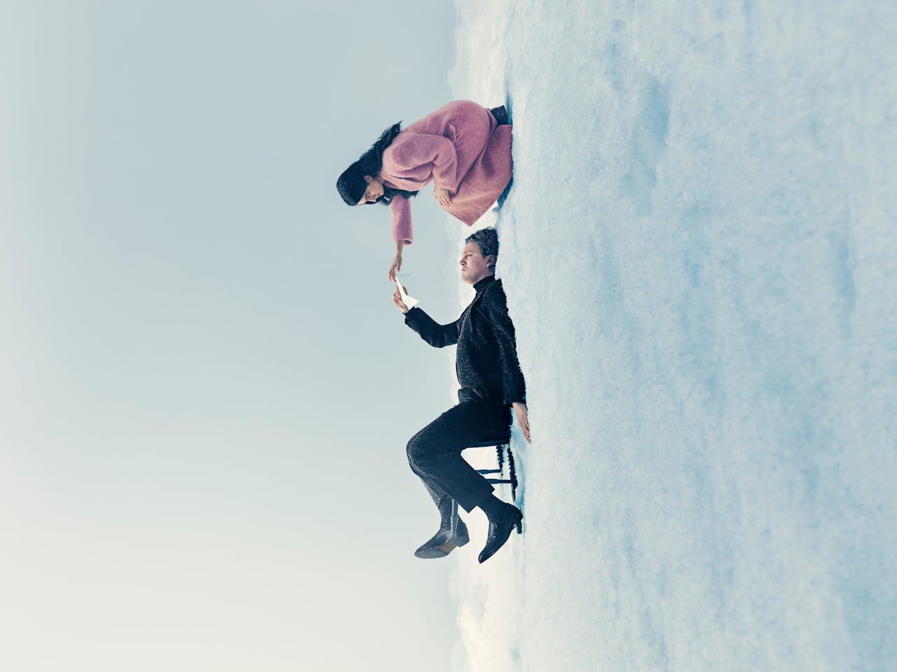 A promotional image for Eugene Onegin. A man dressed in a dark suit sits on a chair on the ground with a woman in pink crouching above him and reaching for his hand.