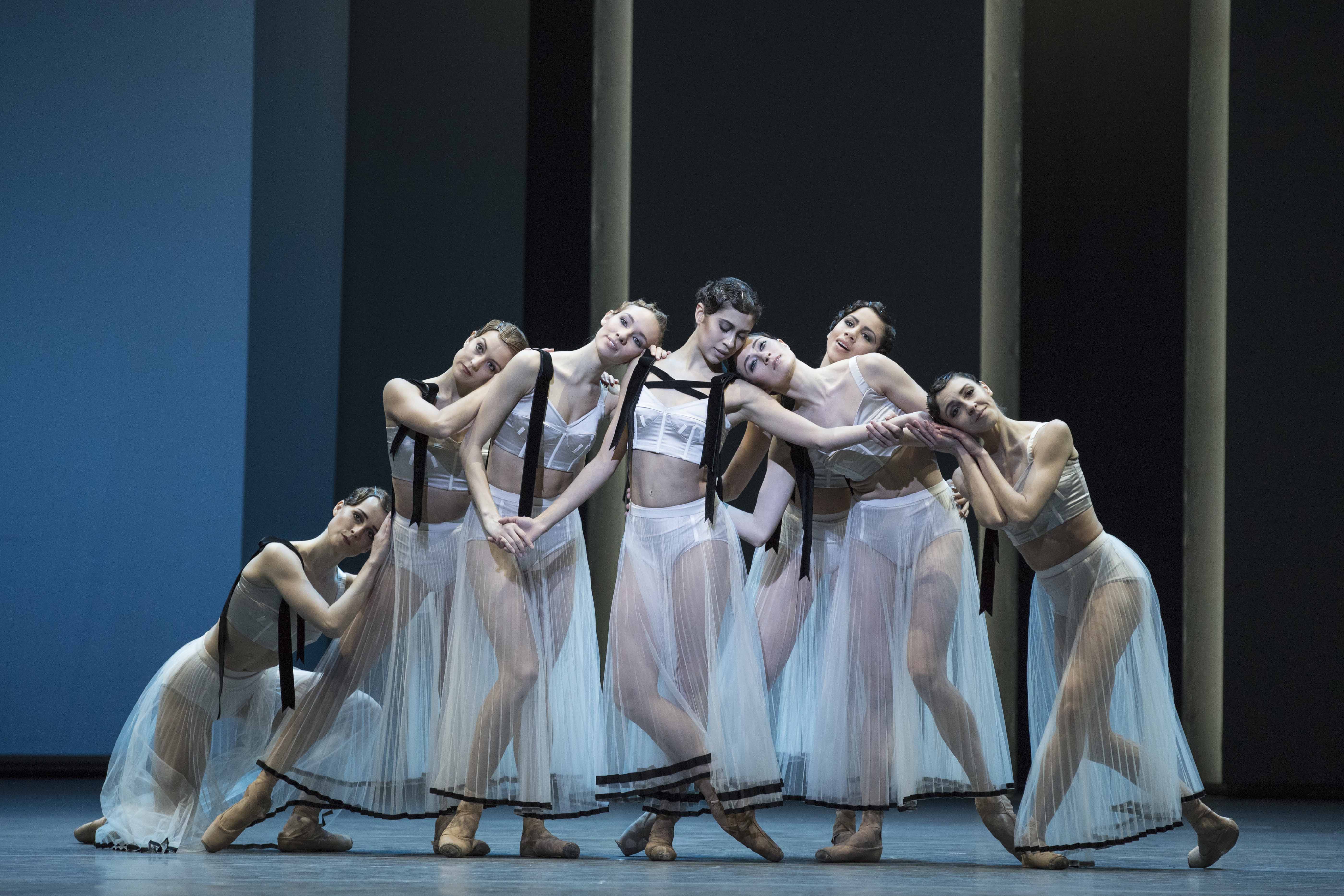 Female dancers in white, sheer costumes stand close together, leaning on each other.