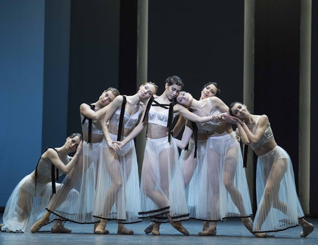 Female dancers in white, sheer costumes stand close together, leaning on each other.