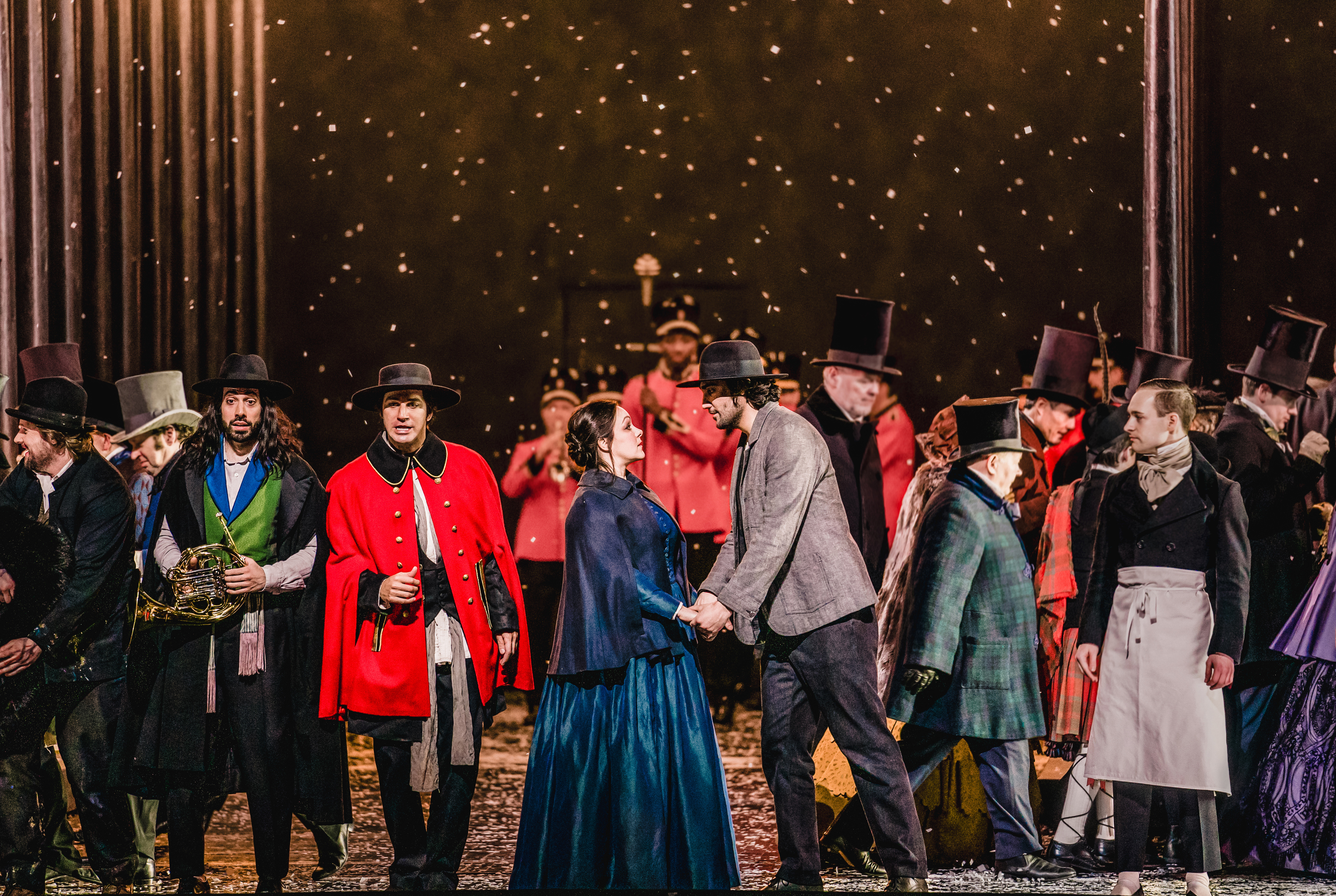 A woman in a blue dress holds hands with a man in a grey suit. They stand on stage in front of a crowd of people dressed in colourful and warm winter clothes. Snow falls from above.