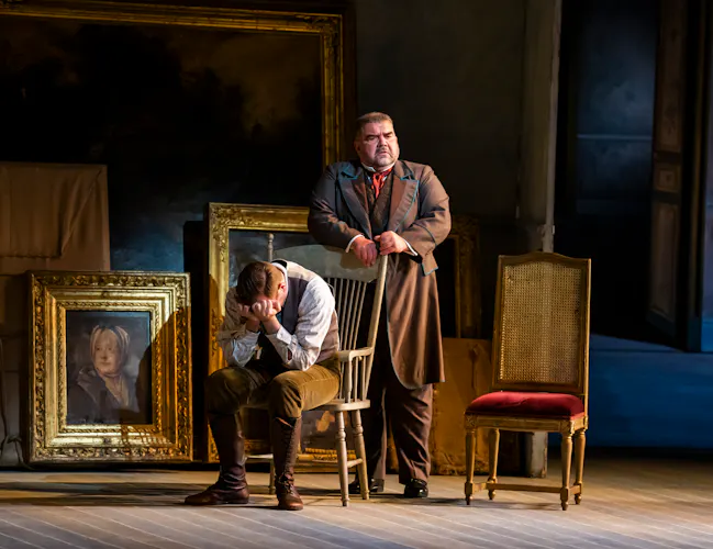 A man sits on a chair with his head in his hands. An older gentleman stands behind him. There are painting in golden frames propped up against the wall in the background.