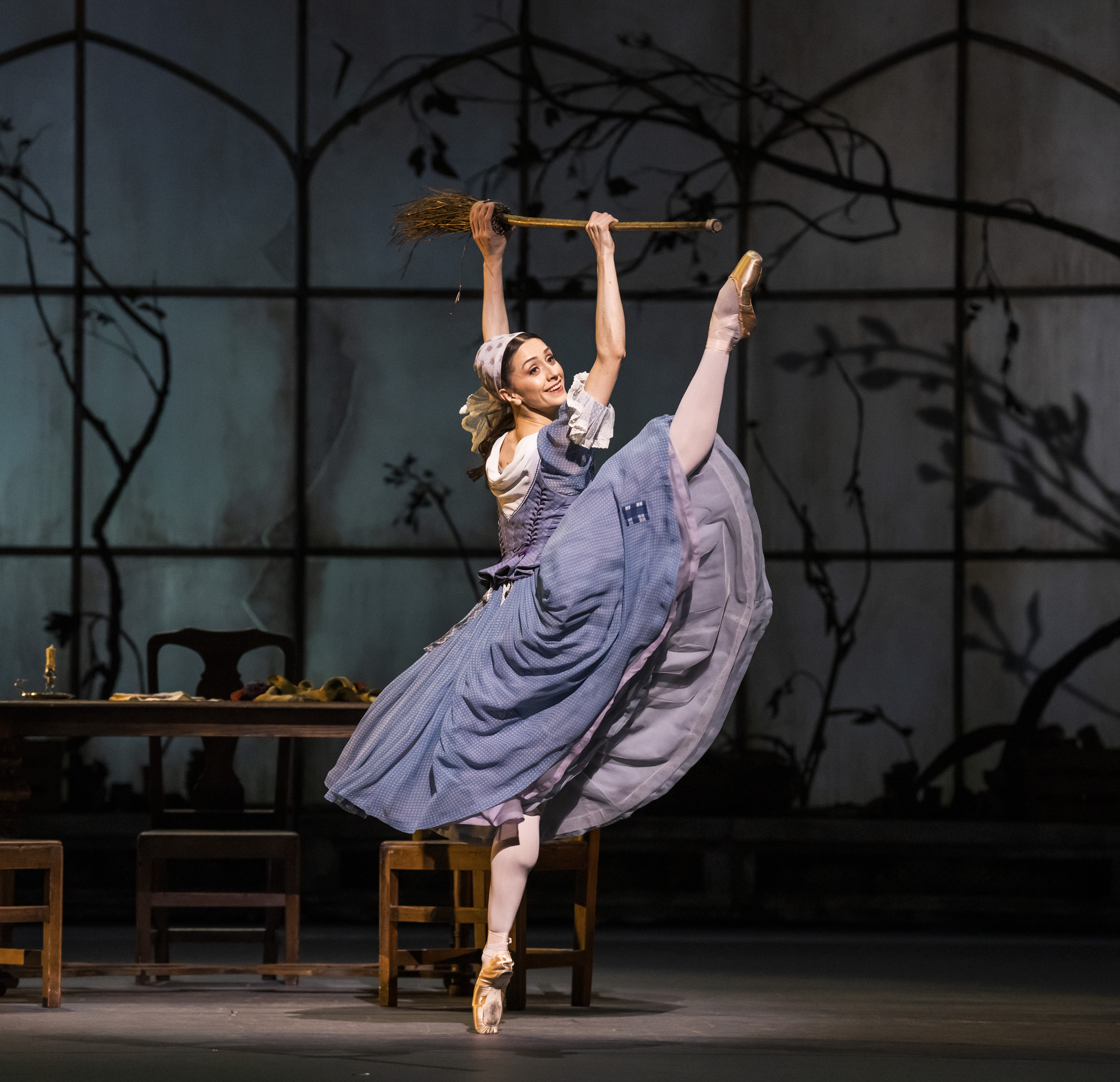 Marianela Nunez performing onstage as Cinderella with a broom. The dancer lifts her leg high in the hair. They are wearing a blue dress and a headscarf. They hold a broom above their head.