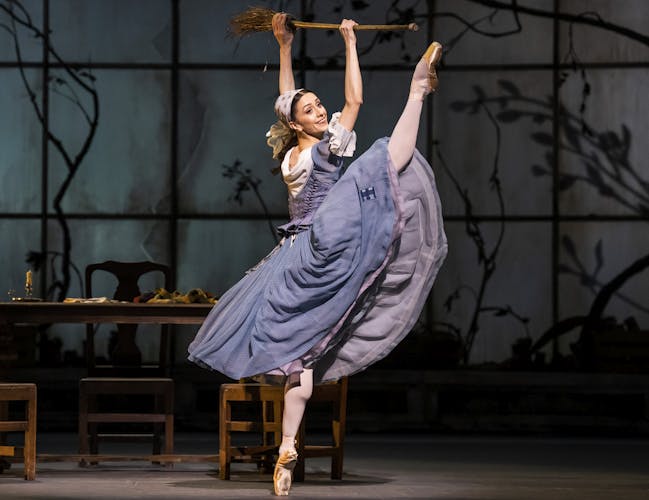 Marianela Nunez performing onstage as Cinderella with a broom. The dancer lifts her leg high in the hair. They are wearing a blue dress and a headscarf. They hold a broom above their head.