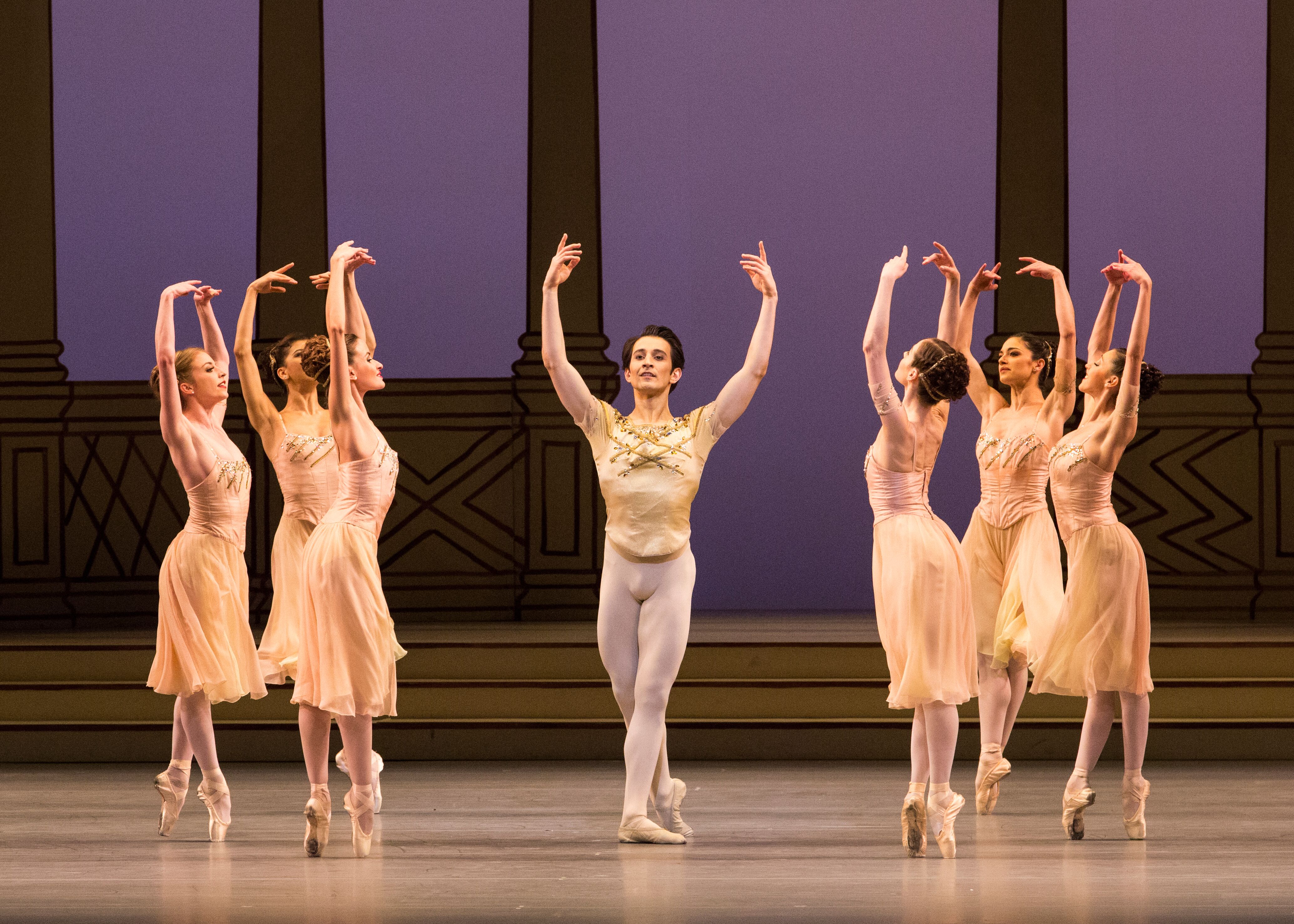 A man in a cream leotard stands in the middle of of a stage. Around him are ballerinas in long cream dresses, balancing on pointe with their arms raised above their heads. (4 of 4)