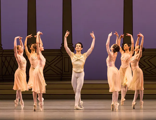 A man in a cream leotard stands in the middle of of a stage. Around him are ballerinas in long cream dresses, balancing on pointe with their arms raised above their heads. (4 of 4)