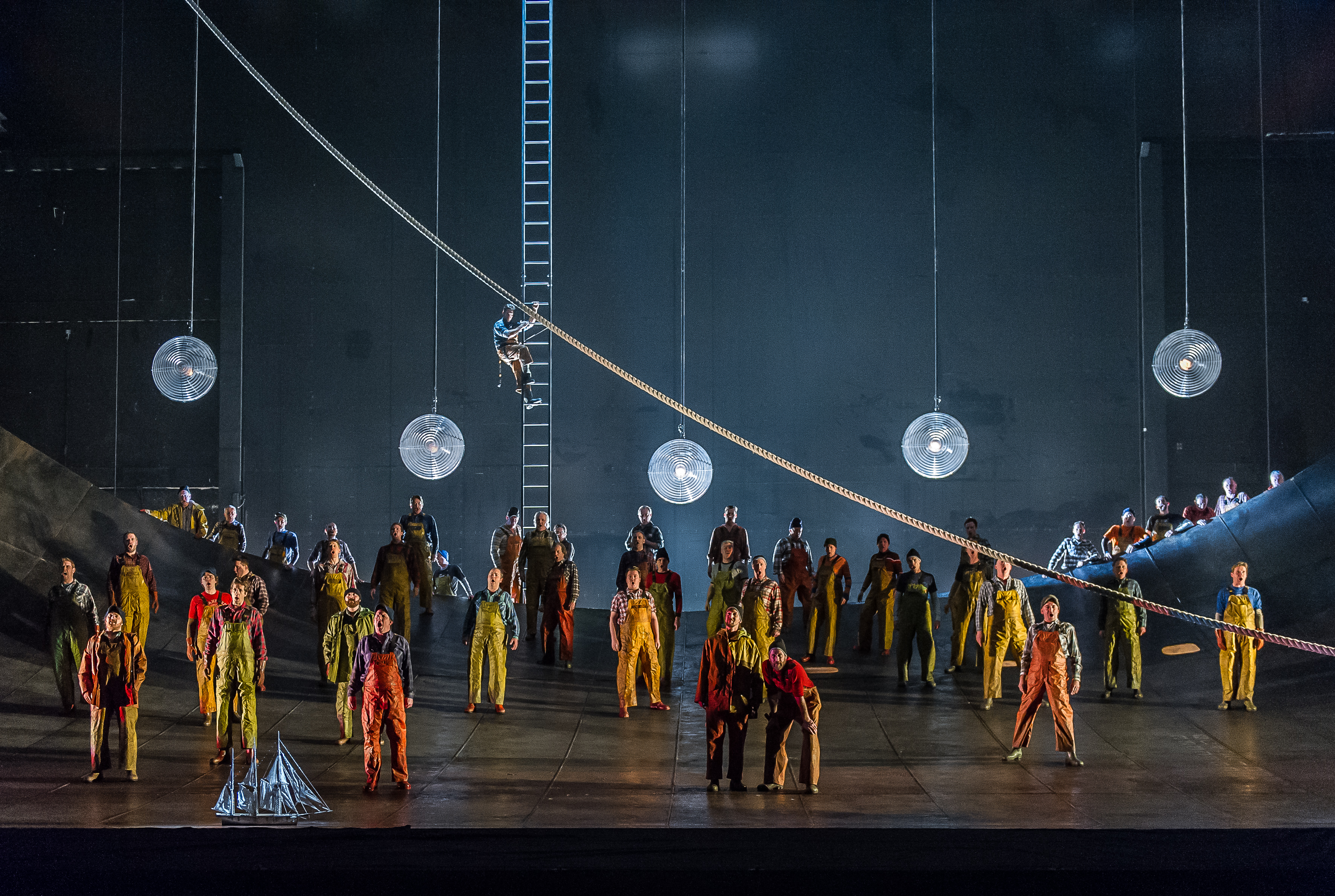 A group of sailors in brightly coloured clothes stand on stage. Large pendant lights hang from the ceiling above the. 
