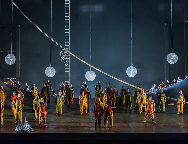 A group of sailors in brightly coloured clothes stand on stage. Large pendant lights hang from the ceiling above the.
