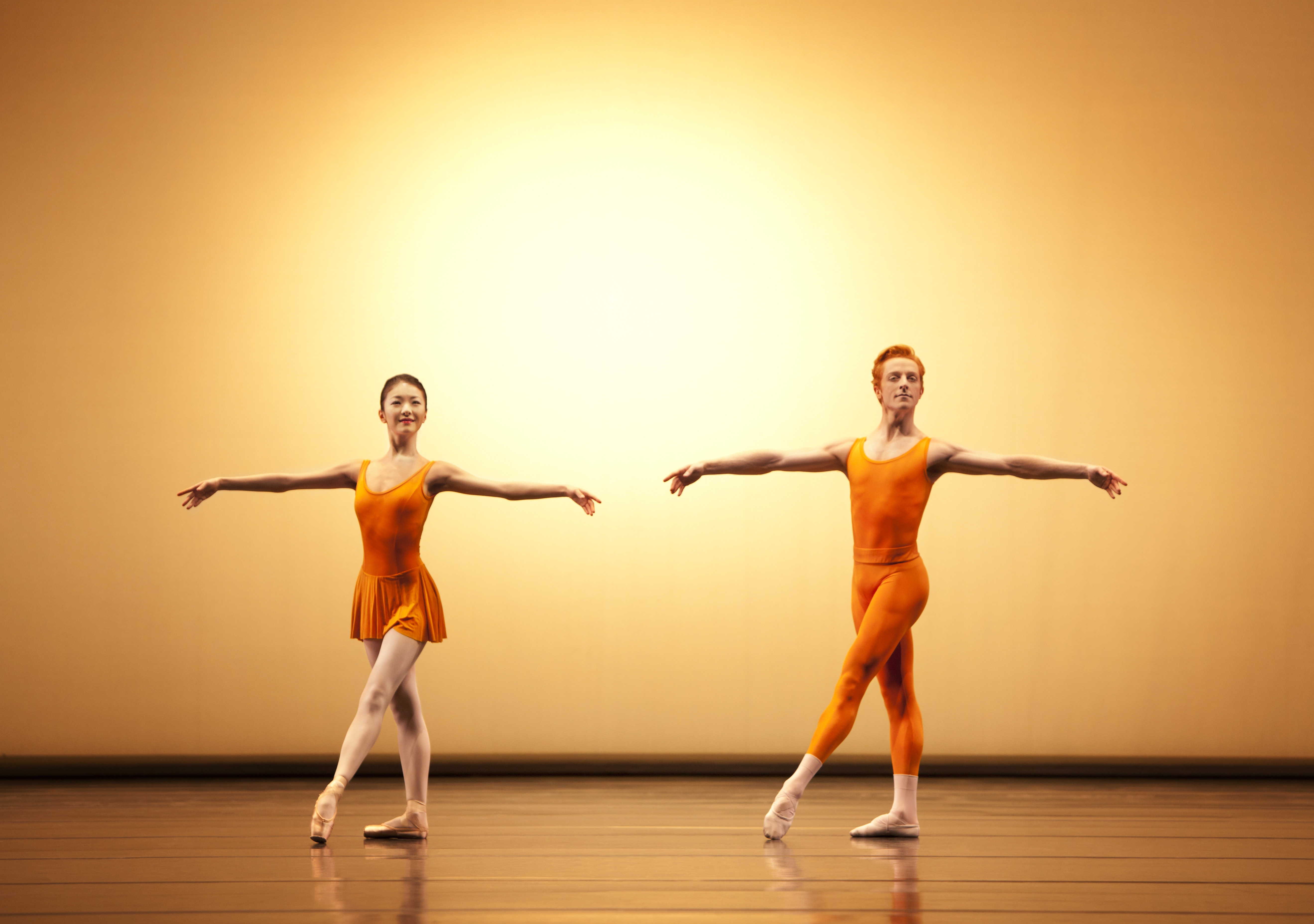 Two dancers wearing yellow leotards appear on stage. The dancer in front crouches with one knee bent in front of him and arms outstretched to the side. Behind him the other dancer has her legs in the splits in a vertical line from the floor, she balances her body on the man's back.