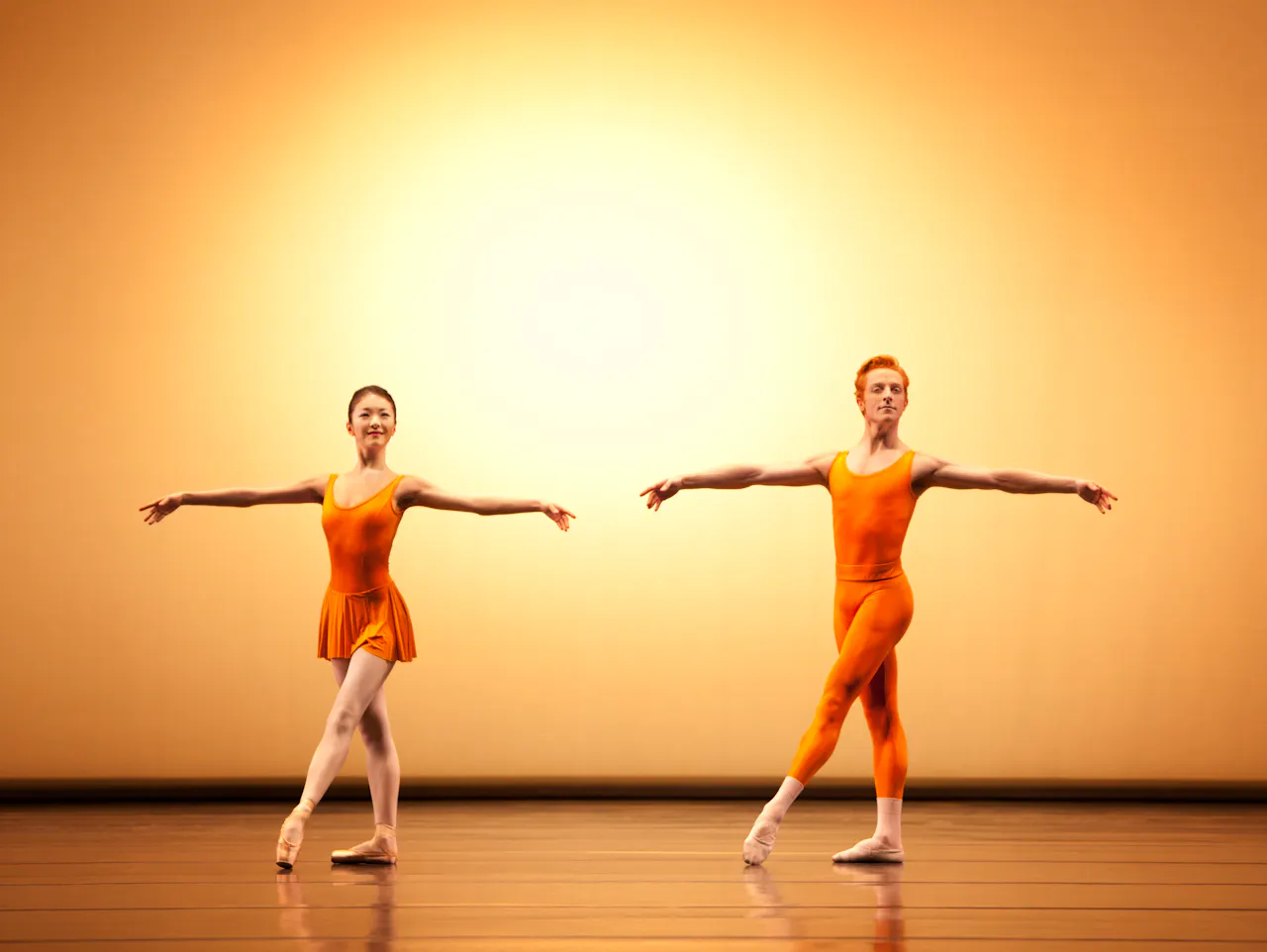 Two dancers wearing yellow leotards appear on stage. The dancer in front crouches with one knee bent in front of him and arms outstretched to the side. Behind him the other dancer has her legs in the splits in a vertical line from the floor, she balances her body on the man's back.