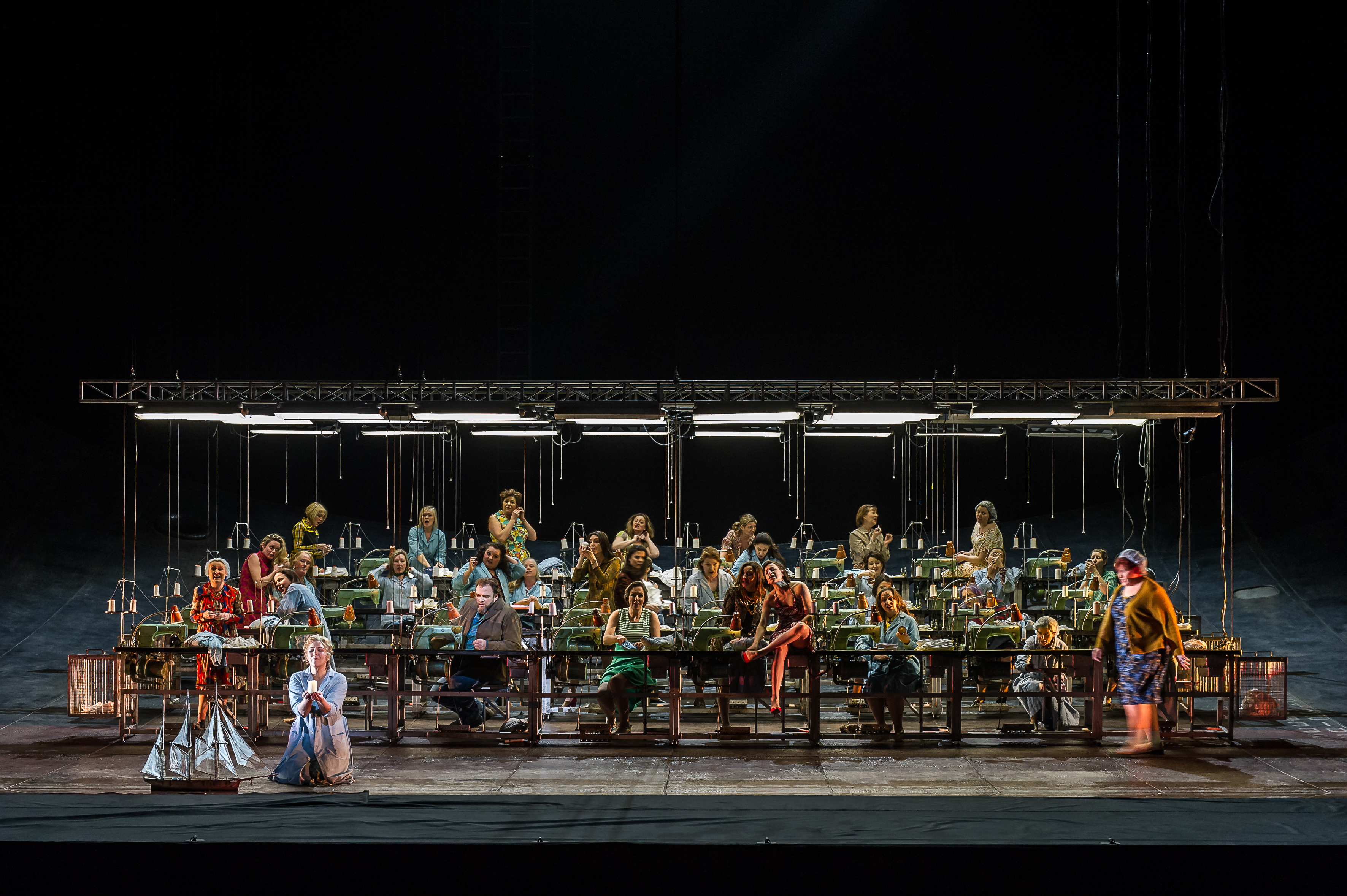 Rows of women sit at industrial sewing machines. A woman kneels at the front next to a model boat. 