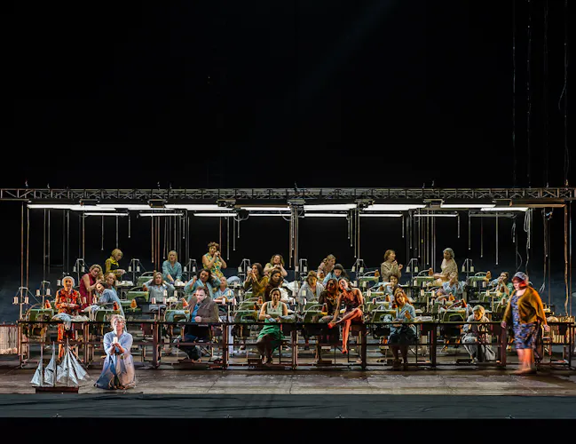 Rows of women sit at industrial sewing machines. A woman kneels at the front next to a model boat.