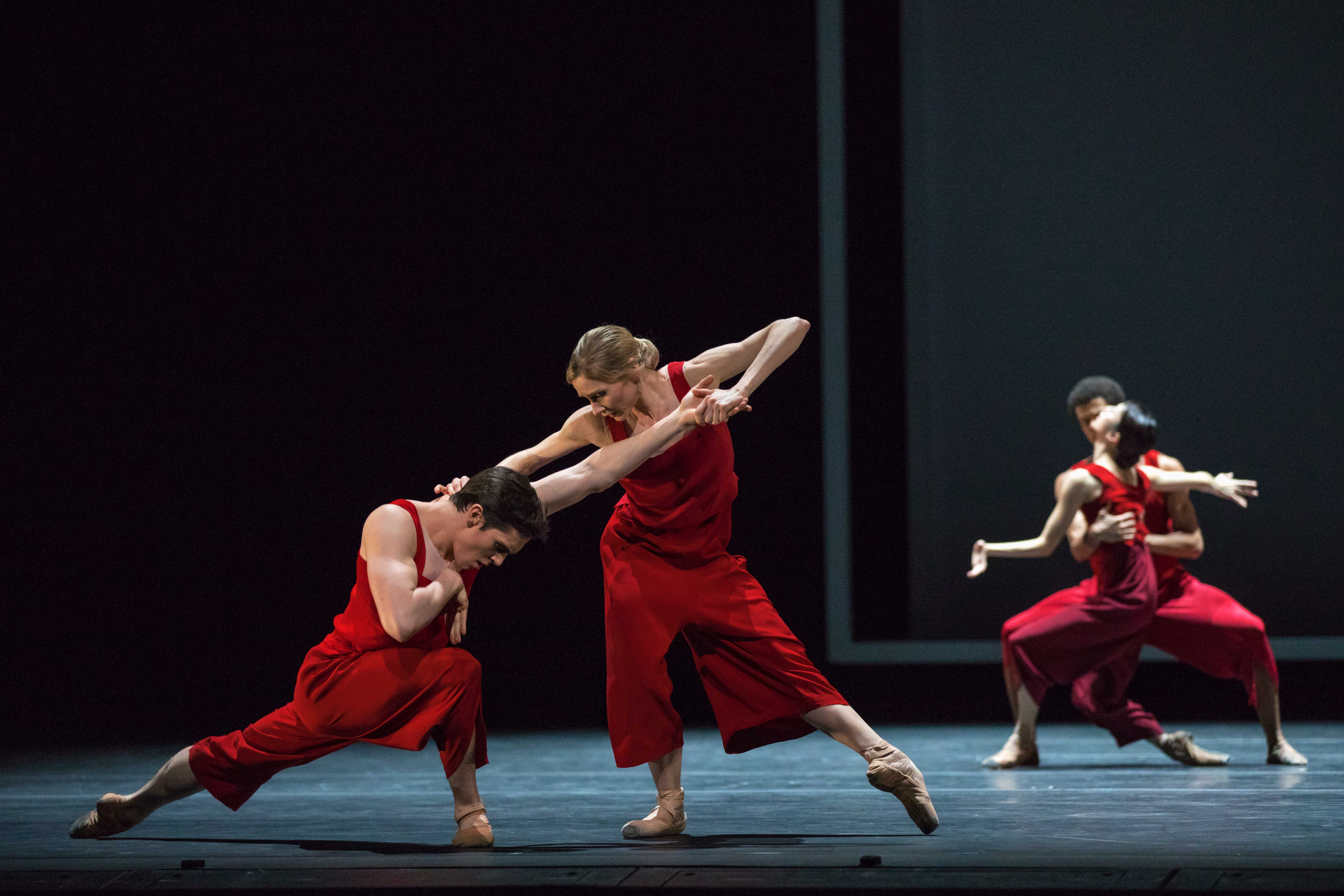 Four dancers in red costumes perform in pairs on a dark, bare stage