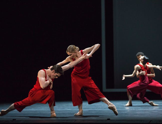 Four dancers in red costumes perform in pairs on a dark, bare stage