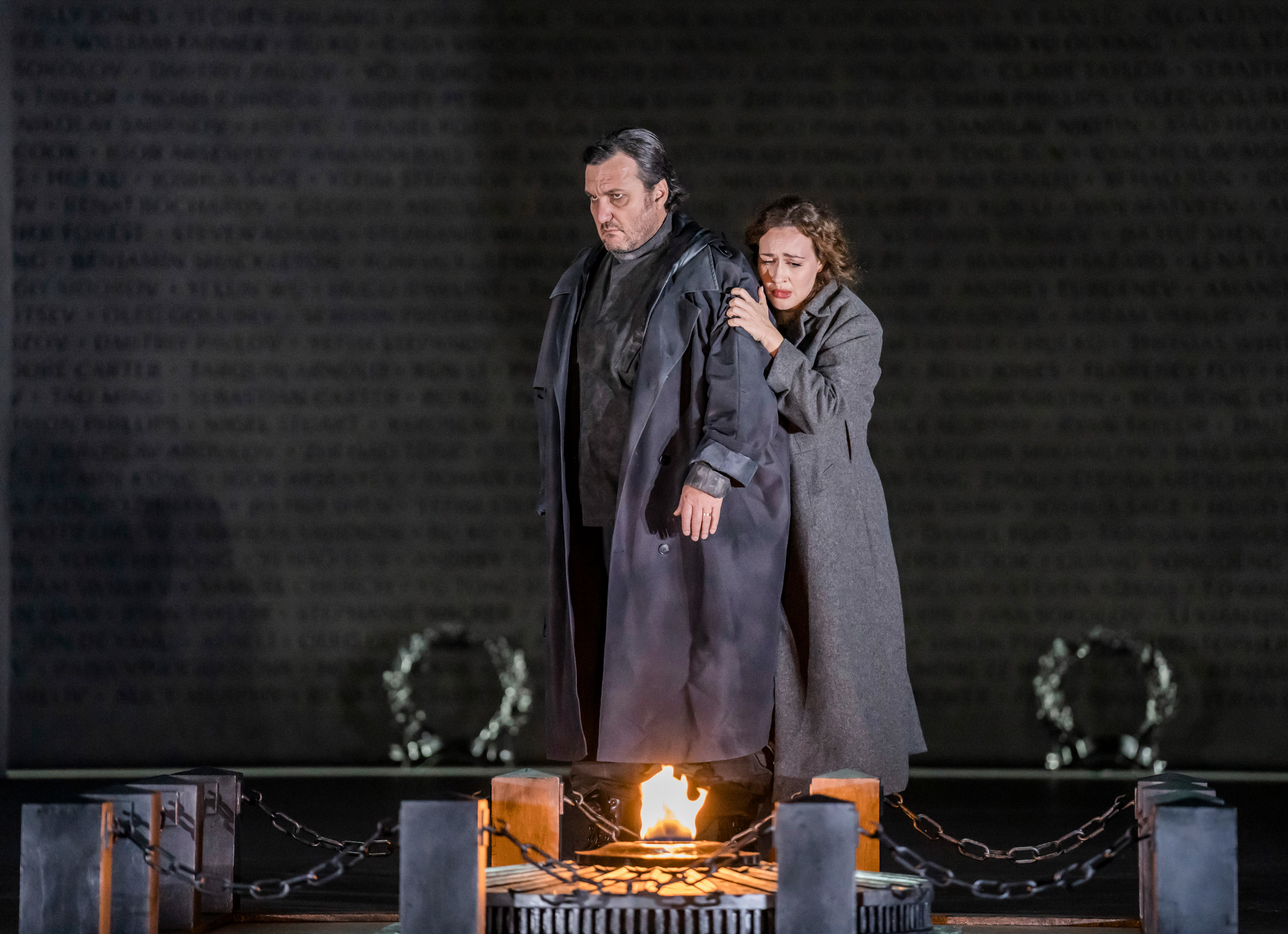 Two characters embrace, one behind the other as they face an open flamed fire. They are both wearing long, heavy grey overcoats
