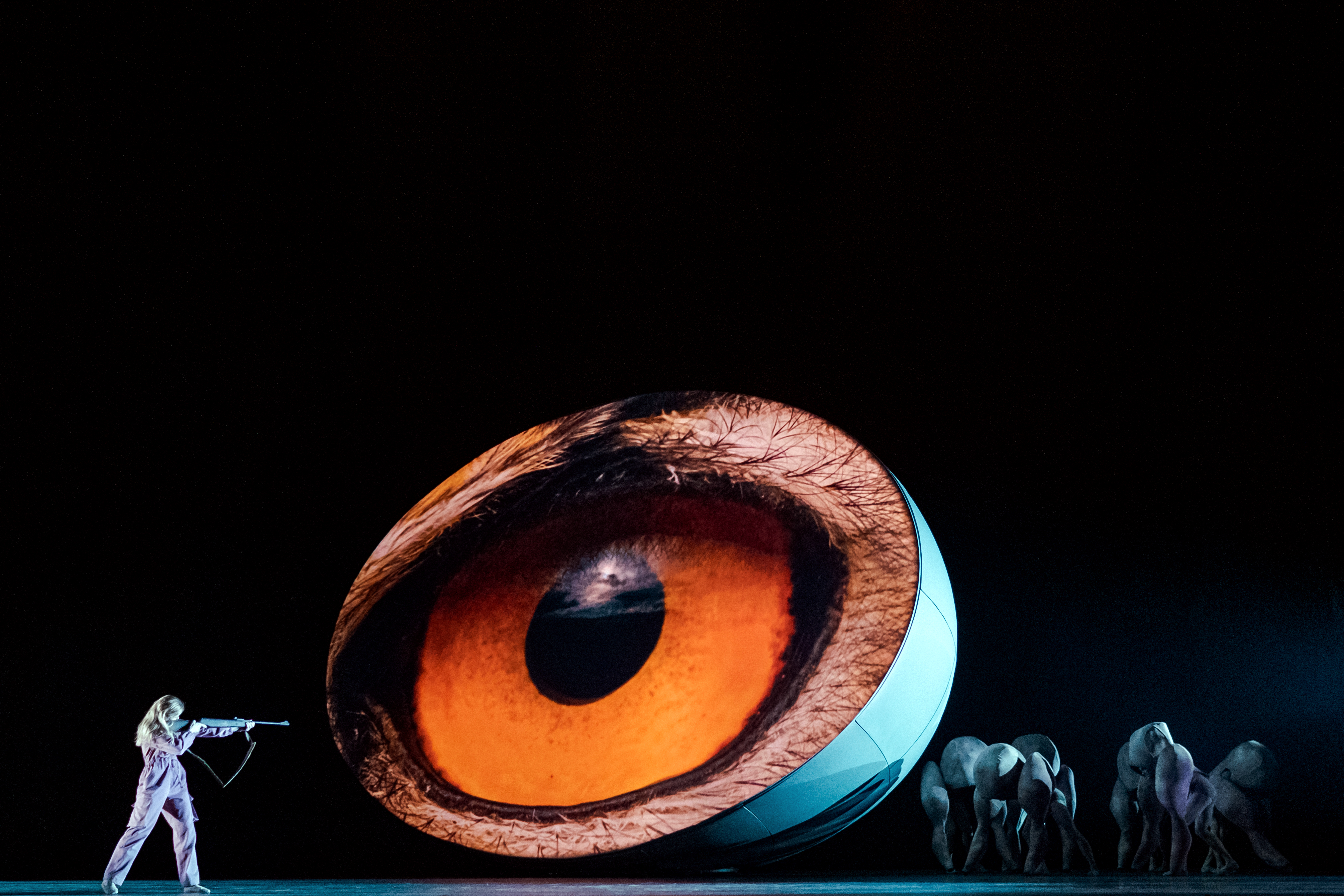 A large orange eye is projected onto a 3d egg shape in the centre of the stage. Alongside, a ballet dancer holds up their arm pointing towards it. There are several other performers dressed in pigoon costumes at the other side of the eye. 