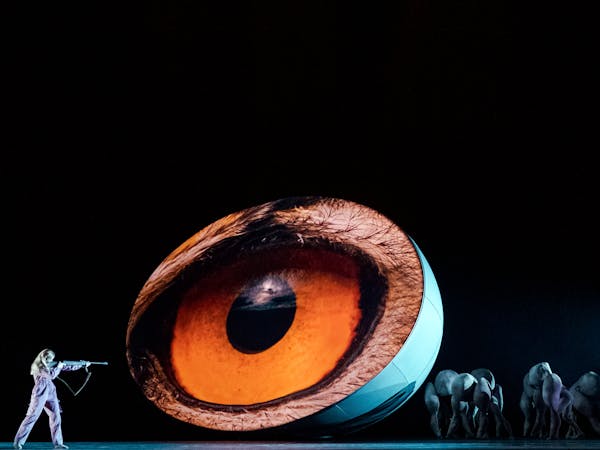 A large orange eye is projected onto a 3d egg shape in the centre of the stage. Alongside, a ballet dancer holds up their arm pointing towards it. There are several other performers dressed in pigoon costumes at the other side of the eye.