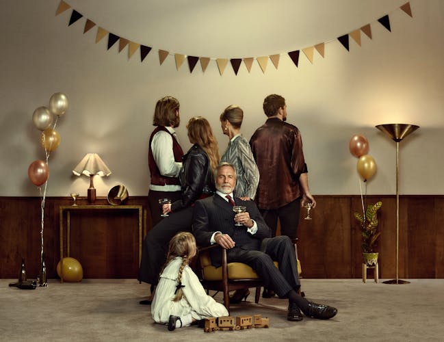 A family in a beige room decorated with bunting and balloons for a celebration. An older man sits on a chair surrounded by his family, who are facing away from the viewer.