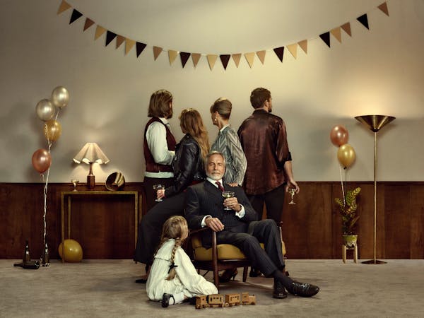 A family in a beige room decorated with bunting and balloons for a celebration. An older man sits on a chair surrounded by his family, who are facing away from the viewer.
