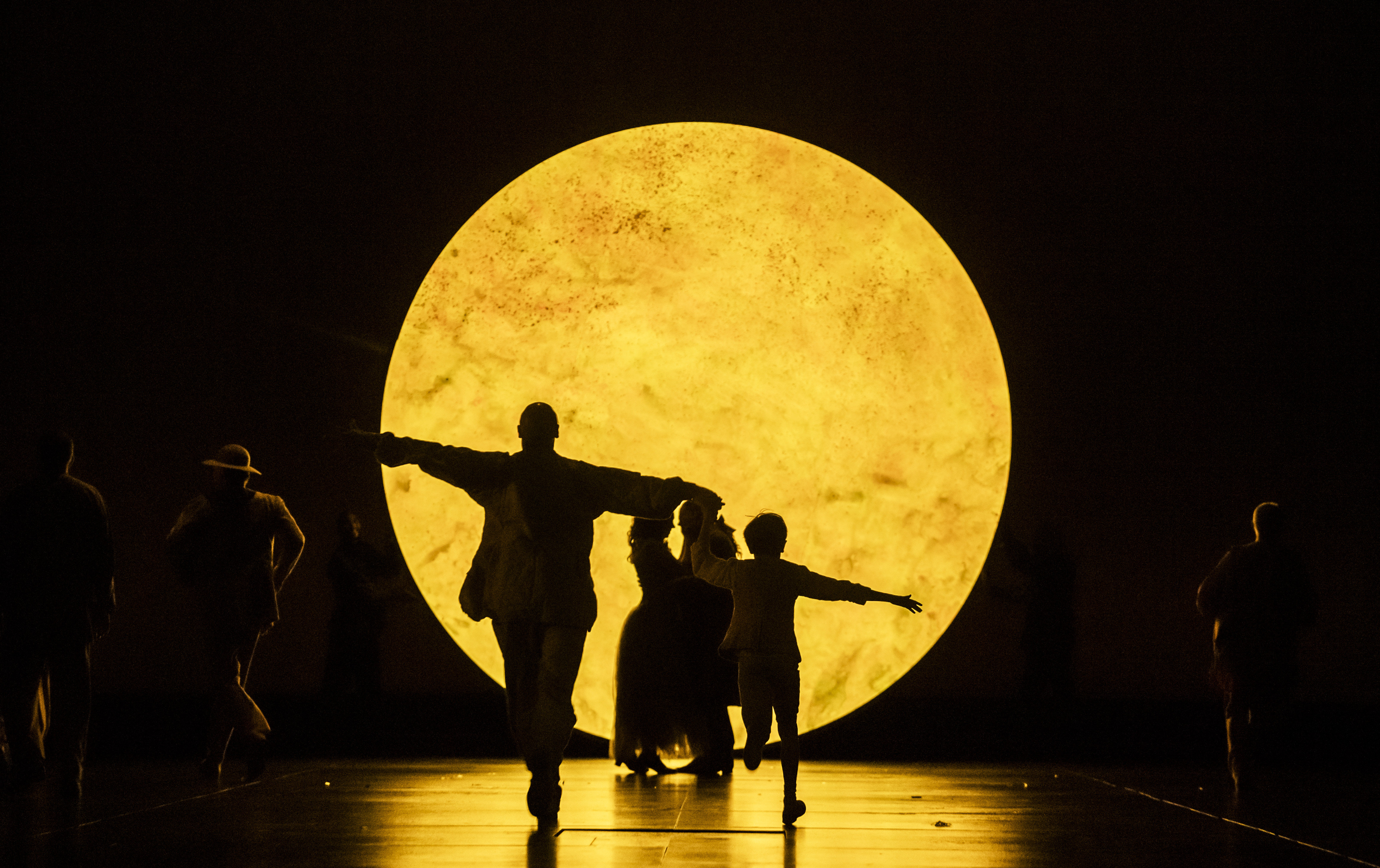Silhouetted people with outstretched arms walk in front of a large, glowing, yellow circular light resembling the moon.