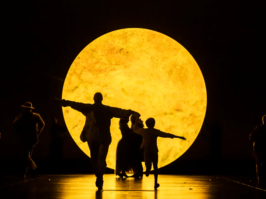 Silhouetted people with outstretched arms walk in front of a large, glowing, yellow circular light resembling the moon.