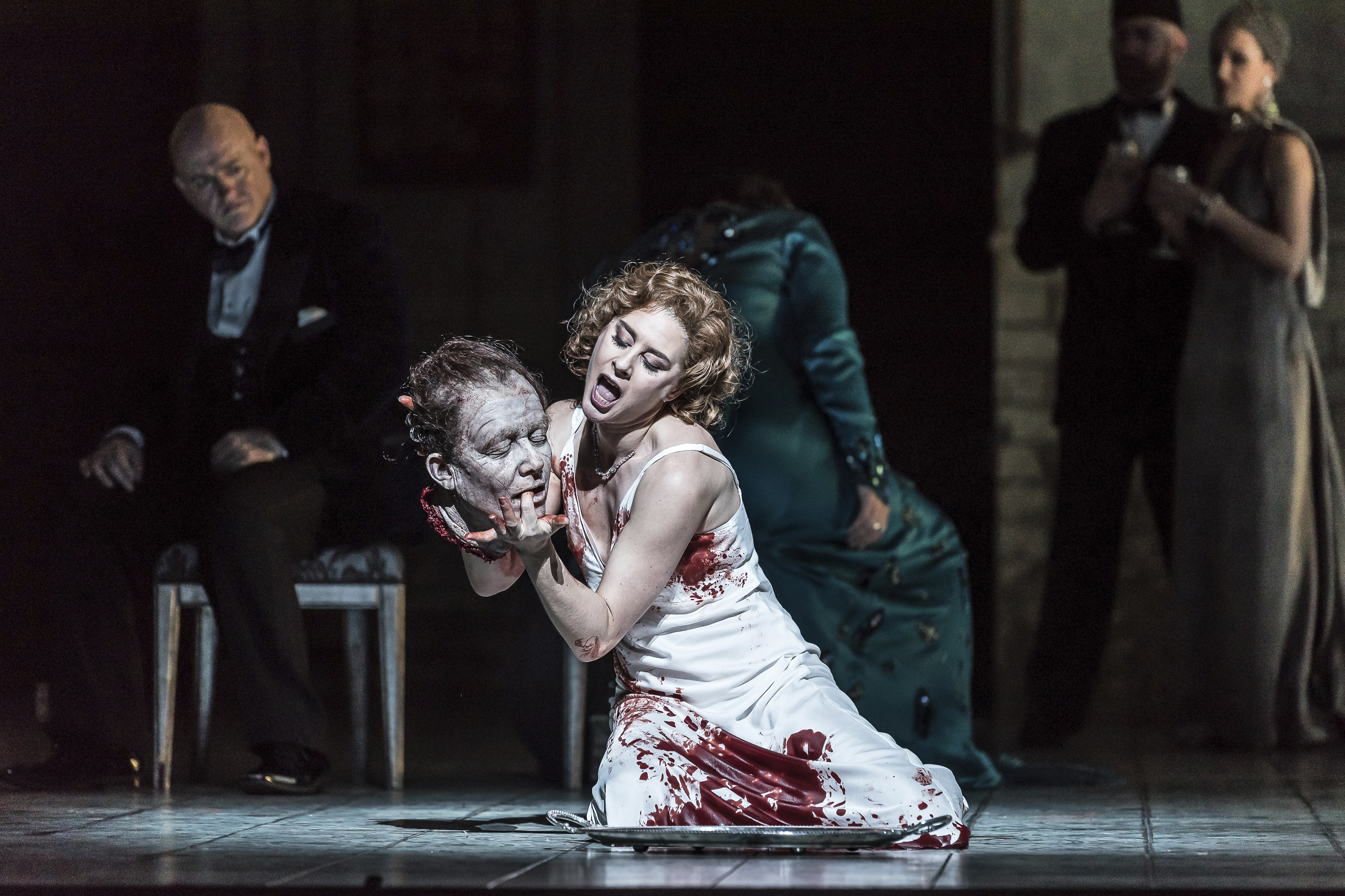 A woman in a blood soaked white dress holds a severed head as she kneels on a stage. 