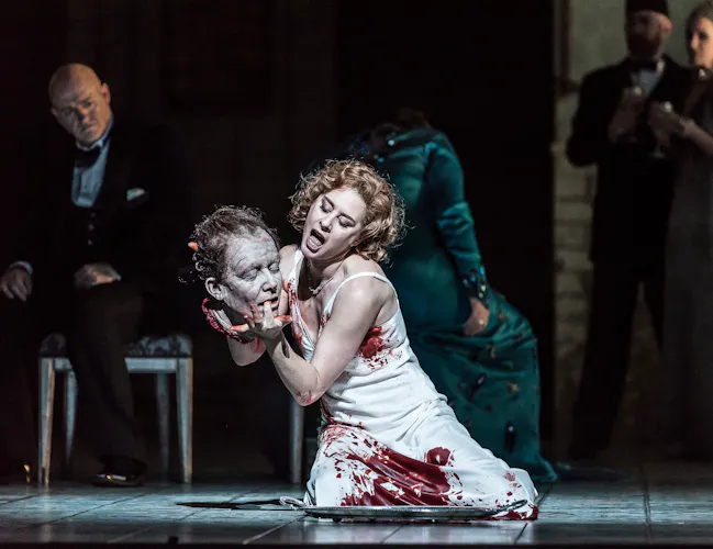 A woman in a blood soaked white dress holds a severed head as she kneels on a stage.