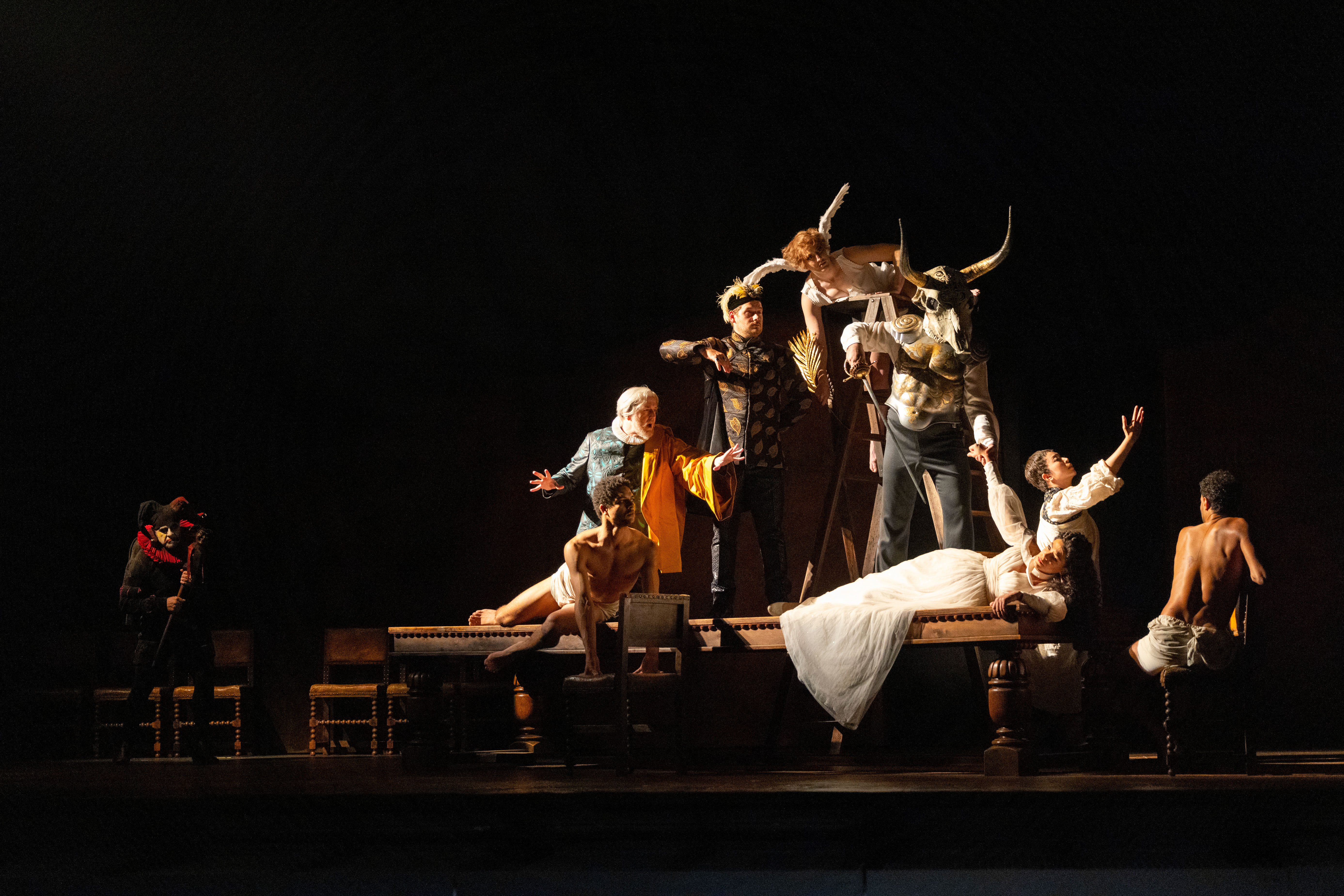 A production image from The Royal Opera's Rigoletto. A character in a white dress holding a glass lays on a table, surrounded by characters in costume.