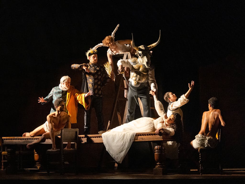 A production image from The Royal Opera's Rigoletto. A character in a white dress holding a glass lays on a table, surrounded by characters in costume.