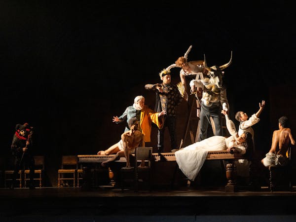 A production image from The Royal Opera's Rigoletto. A character in a white dress holding a glass lays on a table, surrounded by characters in costume.