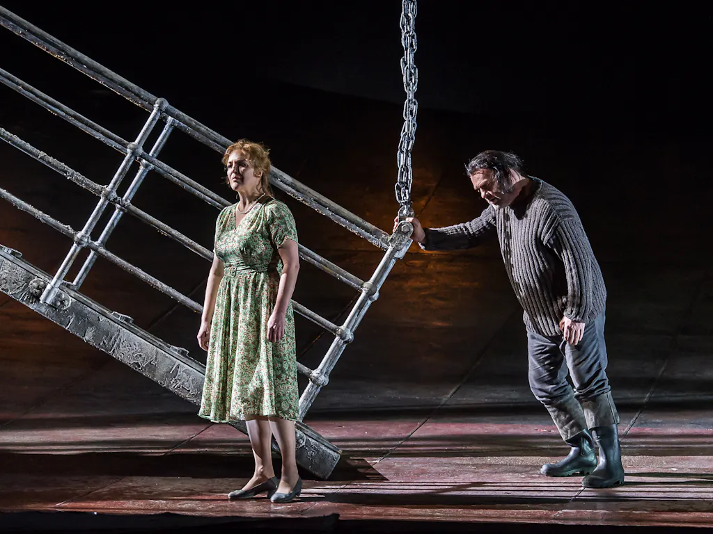 Bryn Terfel as The Dutchman leans on the railing of a metal gangway of a ship. He is looking towards a woman in a green dress who stands looking off into the distance.