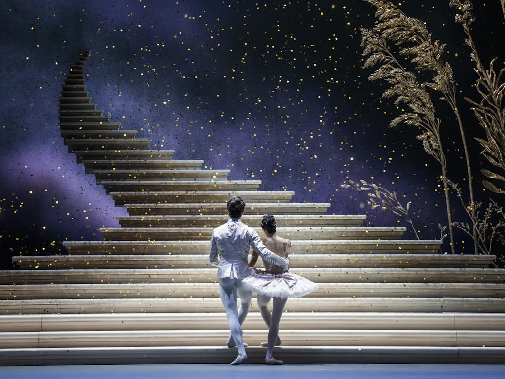 A production of Cinderella performed onstage by the Royal Ballet. The back of a man and woman dressed in white ballet costumes as they look upon a golden staircase that ascends into the sky as glitter falls around them.