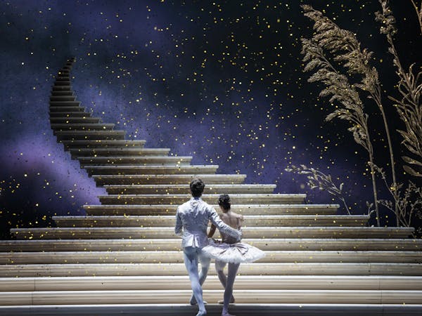 A production of Cinderella performed onstage by the Royal Ballet. The back of a man and woman dressed in white ballet costumes as they look upon a golden staircase that ascends into the sky as glitter falls around them.