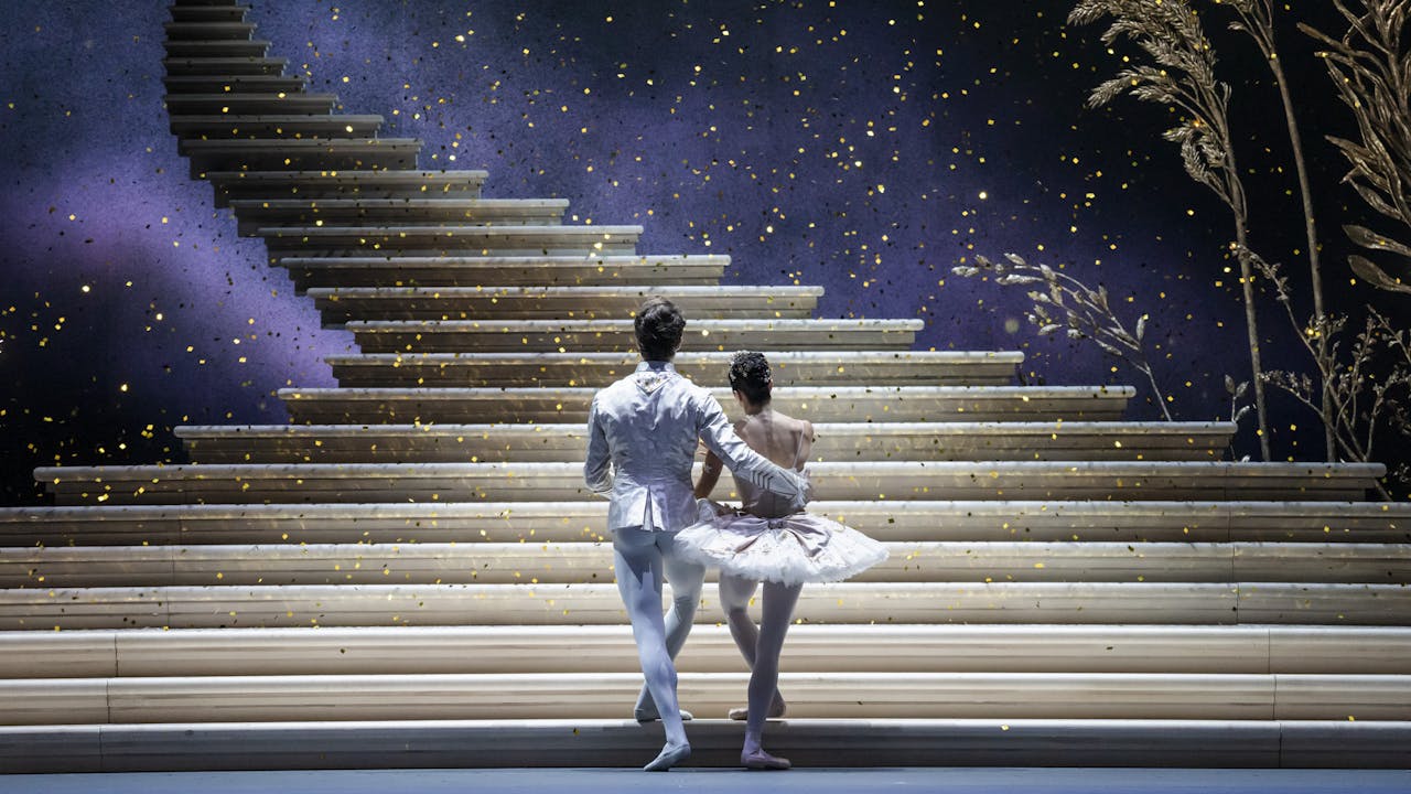 A production of Cinderella performed onstage by the Royal Ballet. The back of a man and woman dressed in white ballet costumes as they look upon a golden staircase that ascends into the sky as glitter falls around them.