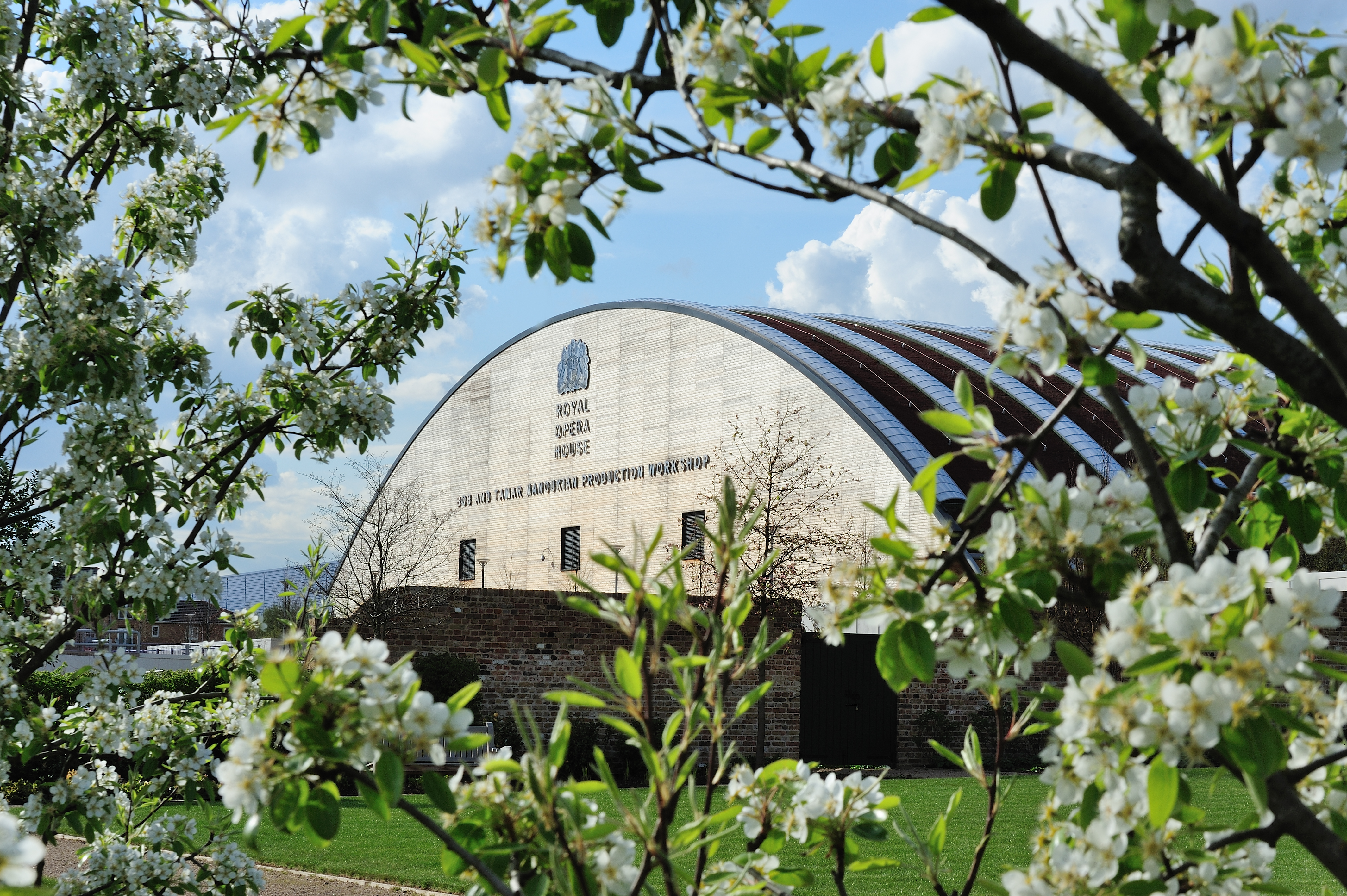 A building with a rounded roof, a brick wall and short-cut grass sits in the foreground and is framed with flowing flowers. 