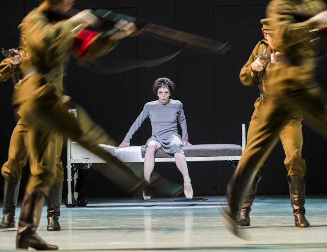 A distressed woman sits on a hospital bed. In front of her, soldiers march.
