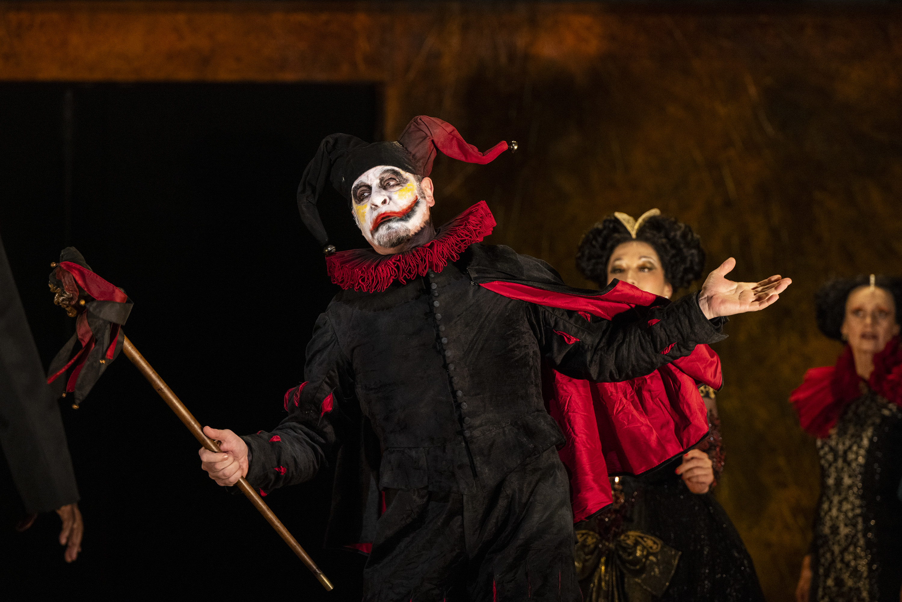 A performer wears a decoratively carved horn skull as a mask and painted steel armour over a white shirt. Their arms are outstretched, and they are holding a sword in their left hand. They are flanked by two women wearing medieval style ballgowns. Behind them are the chorus, all wearing medieval-style clothing. 