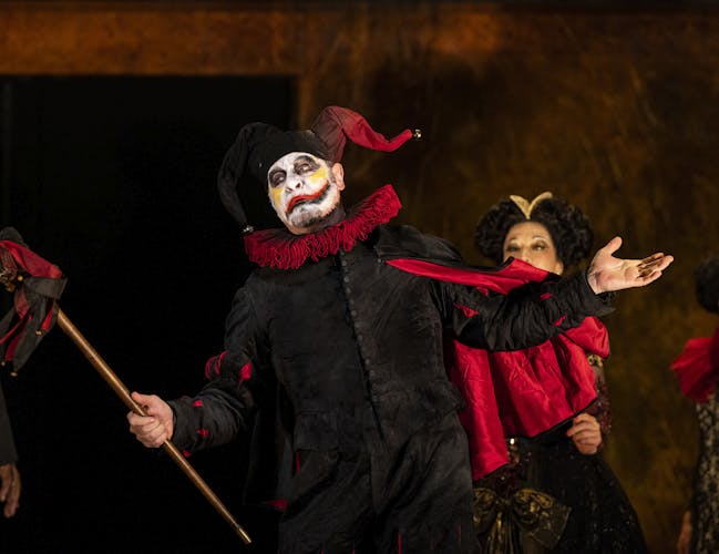 A performer wears a decoratively carved horn skull as a mask and painted steel armour over a white shirt. Their arms are outstretched, and they are holding a sword in their left hand. They are flanked by two women wearing medieval style ballgowns. Behind them are the chorus, all wearing medieval-style clothing.