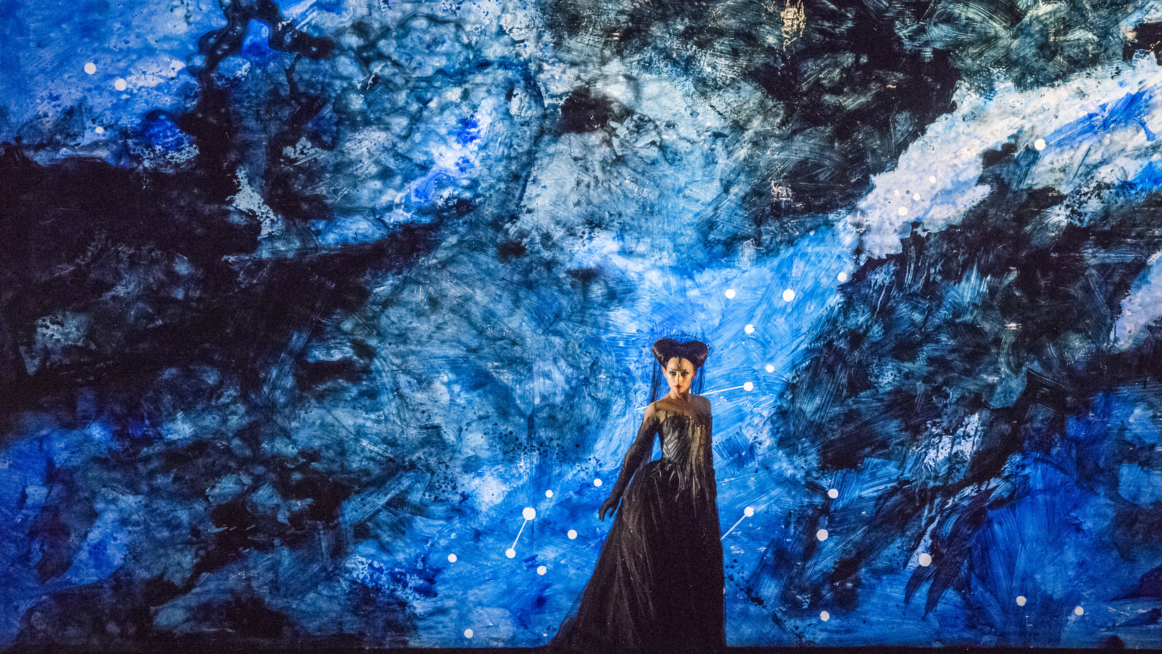 The opera singer Sabine Devieilhe performs as Queen of the Night in The Royal Opera's The Magic Flute. She stands on a stage with a blue and black abstracted starry background wearing a long black gown and her hair is created to look like horns. 