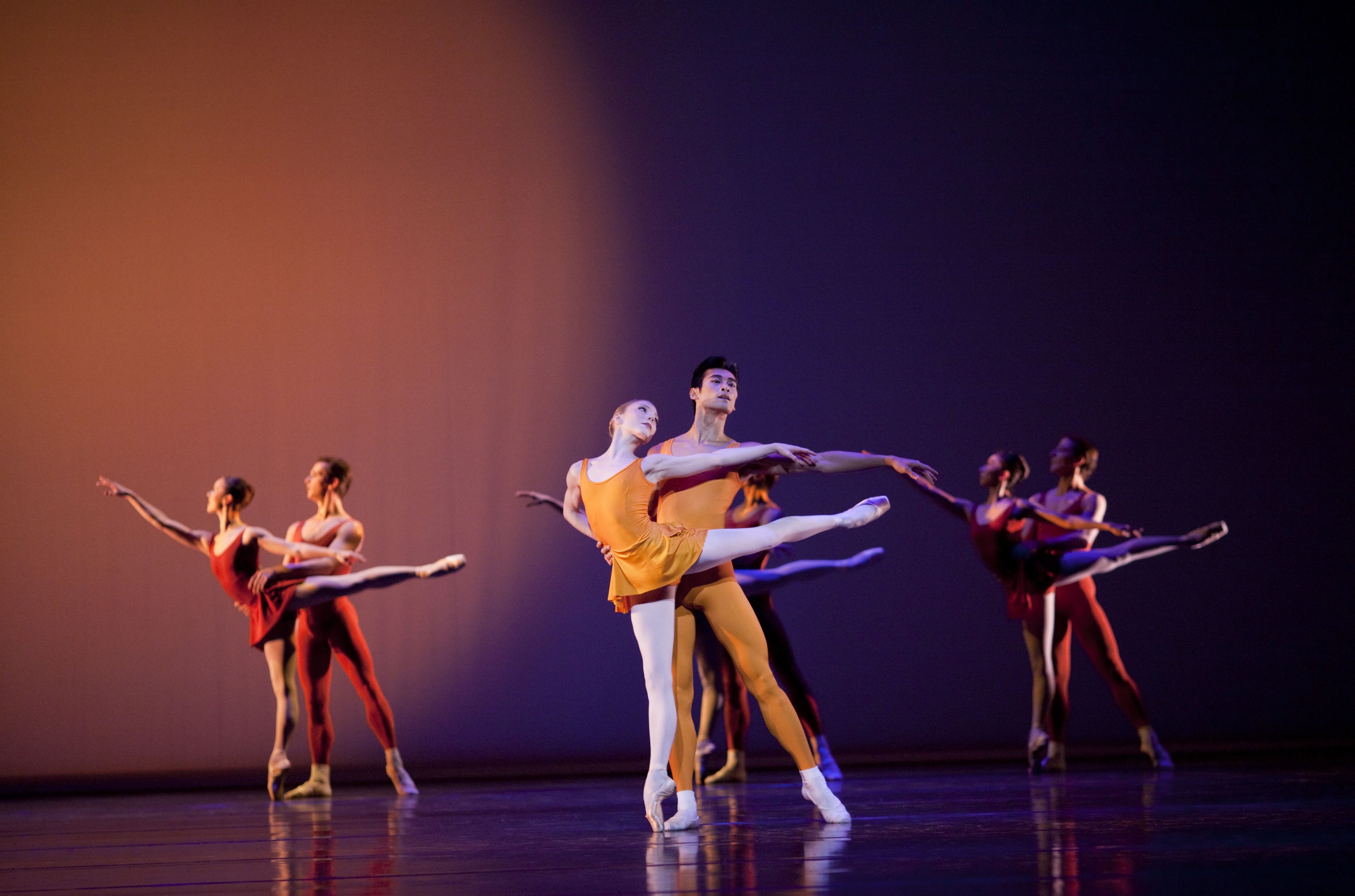 Two dancers in orange leotards perform an arabesque in the foreground. Two couples of dancers do the same in the background wearing red leotards.