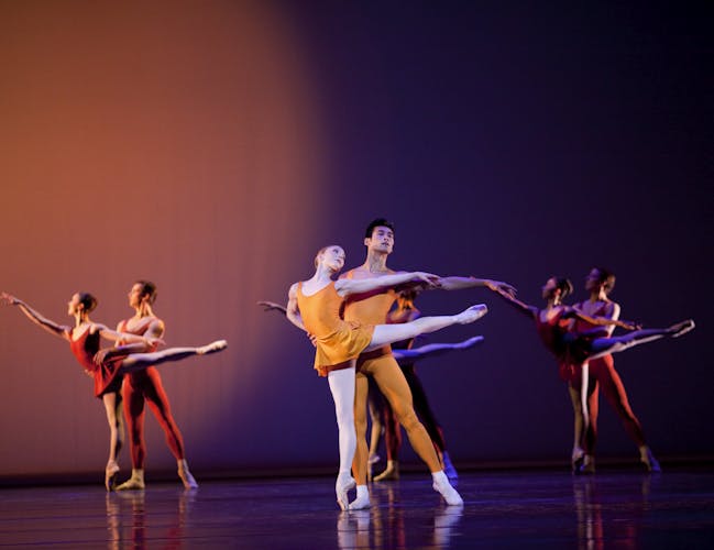 Two dancers in orange leotards perform an arabesque in the foreground. Two couples of dancers do the same in the background wearing red leotards.