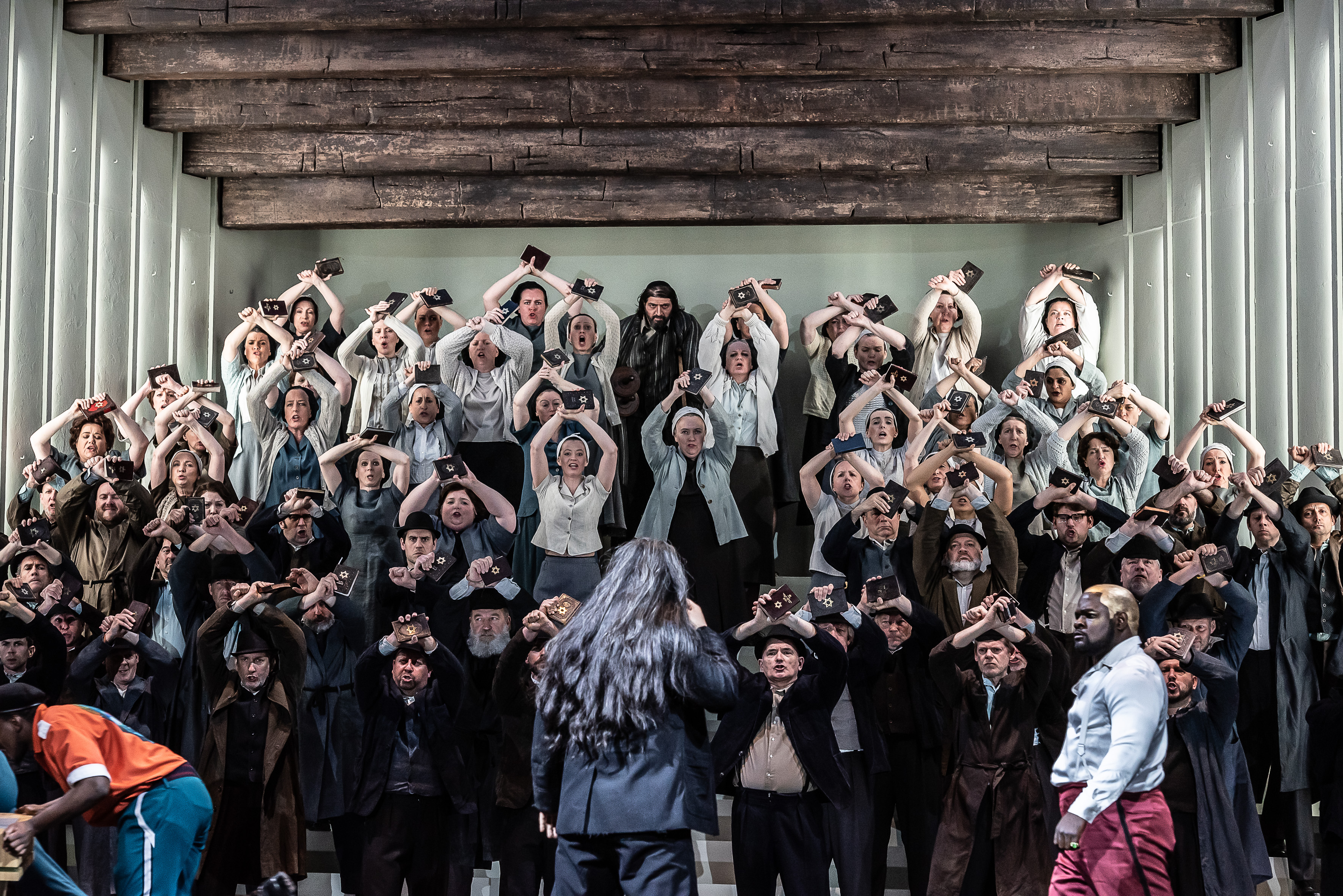 A chorus of singers stand on a stairs on a stage with their arms raised in the air, they are holding up a book with has a gold star embossed on the front. At the centre front, a character wearing red trousers lunges forward holding their chest. Behind them on the stairs leading up are two others, one has their right arm in the air holding up what looks like a knife. The character behind them leans on a crutch.