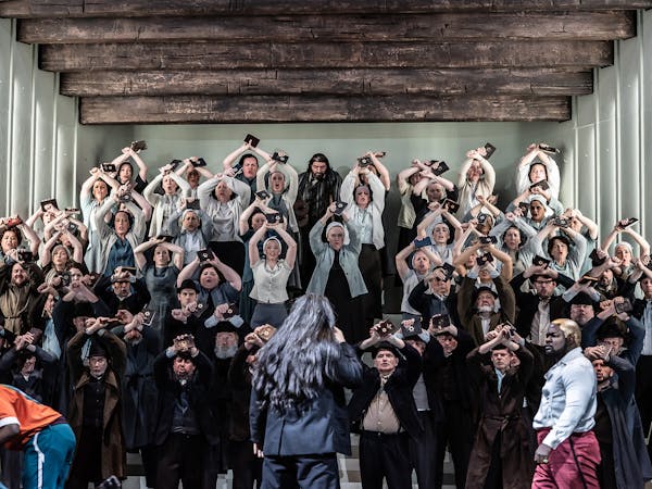 A chorus of singers stand on a stairs on a stage with their arms raised in the air, they are holding up a book with has a gold star embossed on the front. At the centre front, a character wearing red trousers lunges forward holding their chest. Behind them on the stairs leading up are two others, one has their right arm in the air holding up what looks like a knife. The character behind them leans on a crutch.