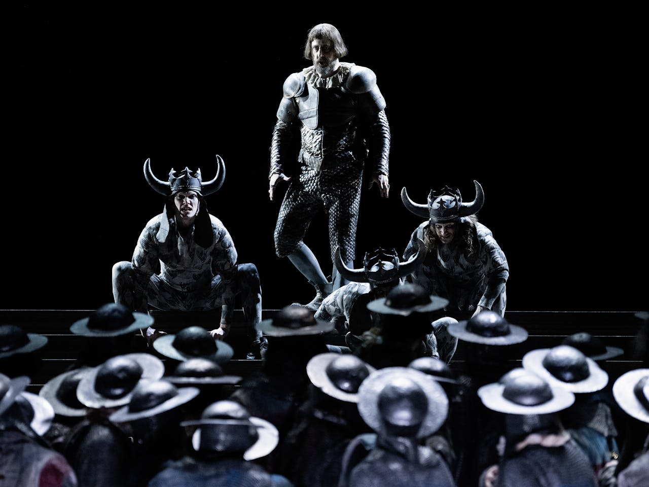 Three actors stand on stage, two are crouched at either side of the central actor and they are wearing horned helmets. They are faced by an army wearing helmets.