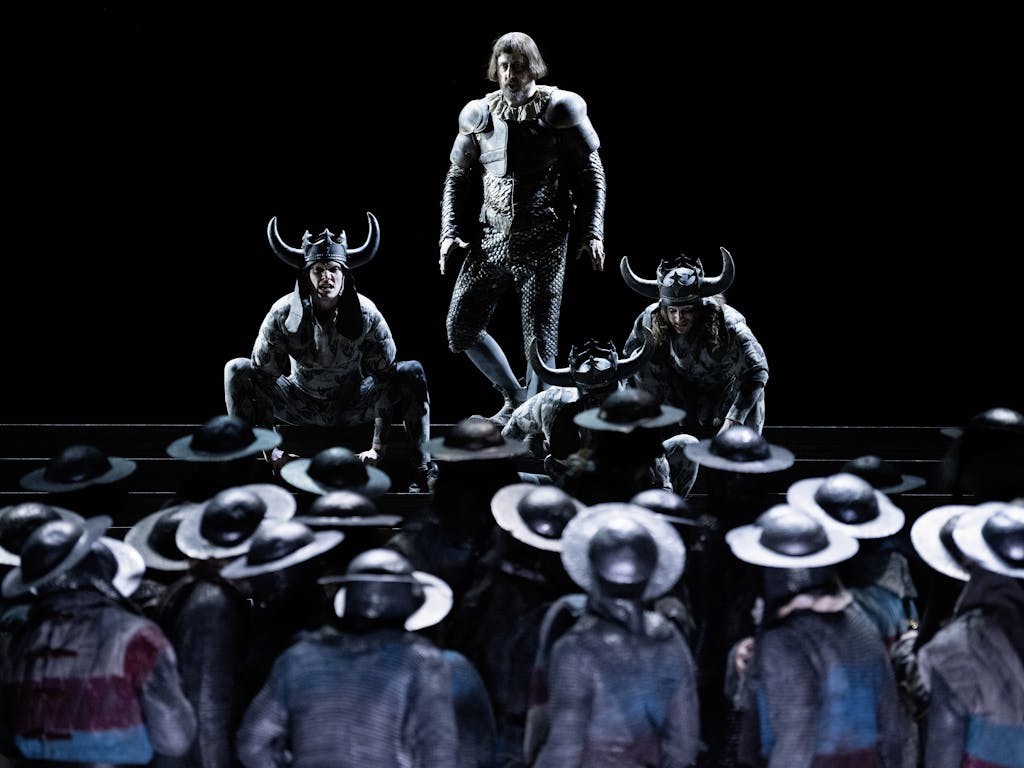 Three actors stand on stage, two are crouched at either side of the central actor and they are wearing horned helmets. They are faced by an army wearing helmets.